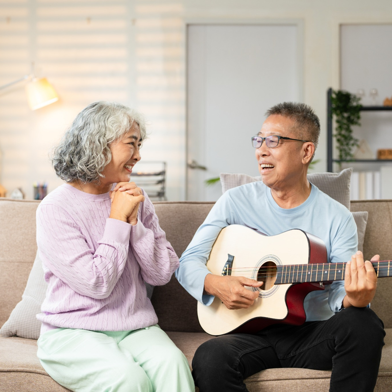 An older couple sits together on a couch at home while one plays the guitar and sings while the other smiles, showing how music-based aphasia practice can support communication, connection, and enjoyment during daily activities.